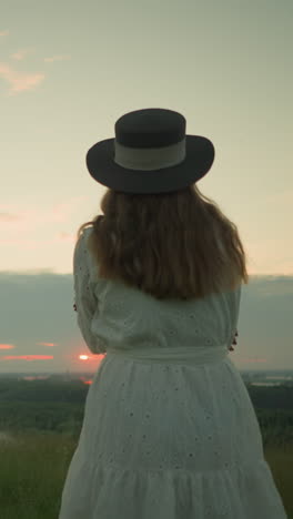 vista de cerca de una mujer con un vestido blanco y sombrero, brazos cruzados, de pie sola en un campo de hierba serena, mirando hacia un lago tranquilo durante la puesta de sol, bajo un cielo suave y de color pastel
