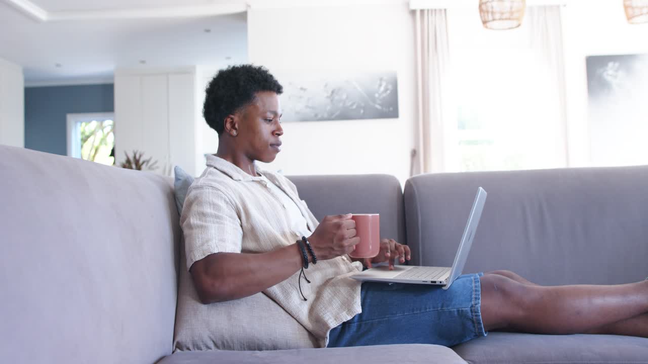 African American man reclining on gray sofa in living room, holding mug and typing laptop