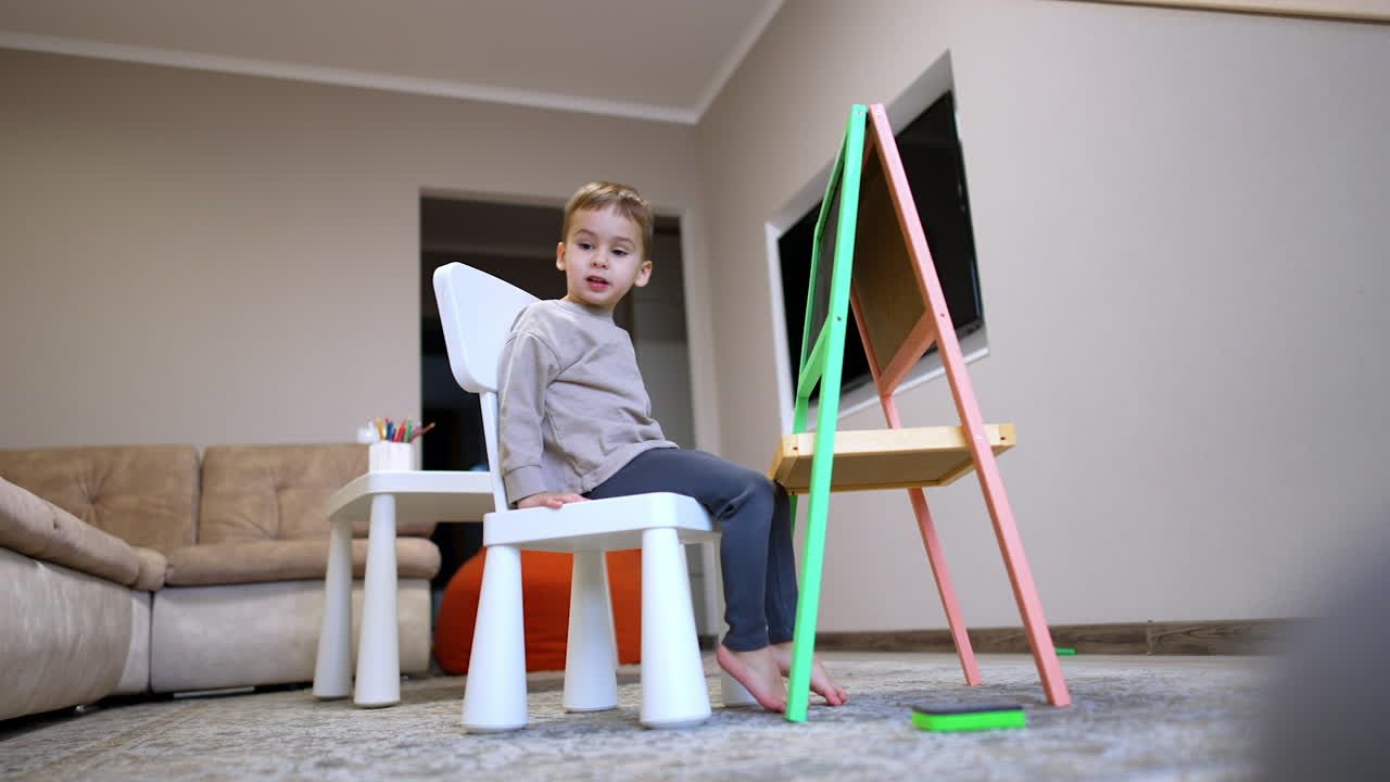 Lovely toddler sits on the chair in front of the blackboard. Low angle view at the kid at home.