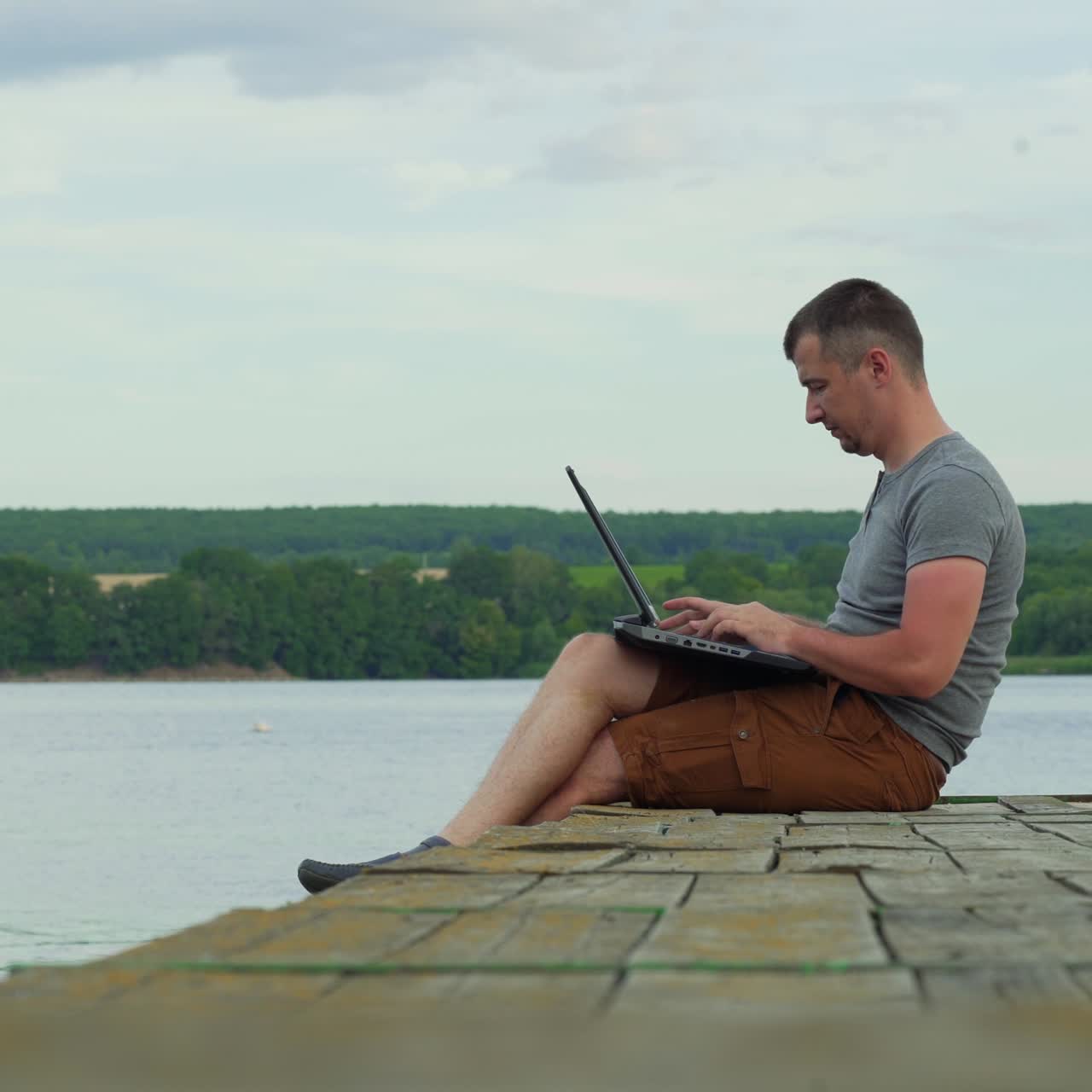 man is sitting on the bridge and opening a laptop to work on the background of the river in the summer