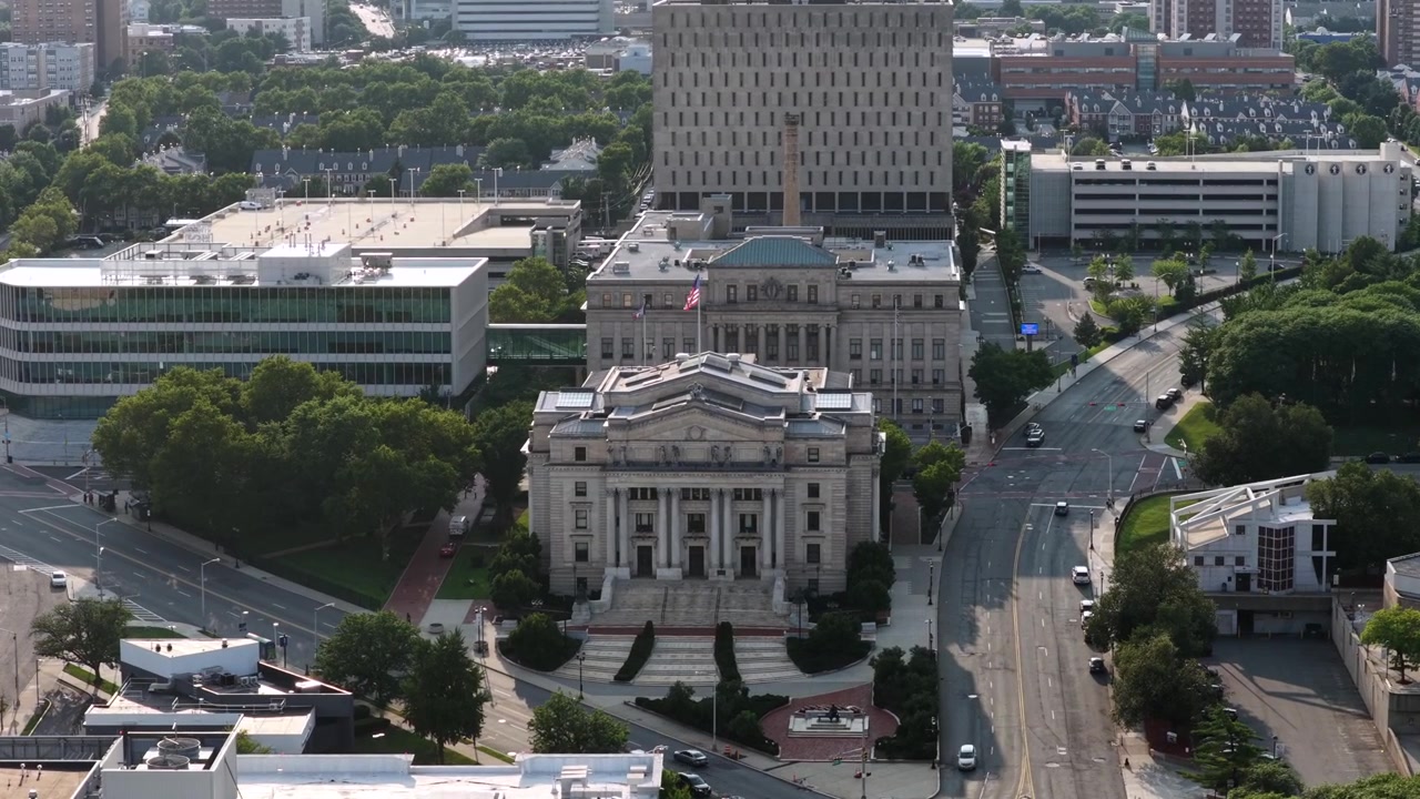 Tribunal com cúpula e colunas clássicas ancora a praça do centro de Newark, com um edifício de escritórios de vidro e um estacionamento emoldurando uma ampla avenida arborizada com tráfego de passagem. Tomada aérea de drone em aproximação.
