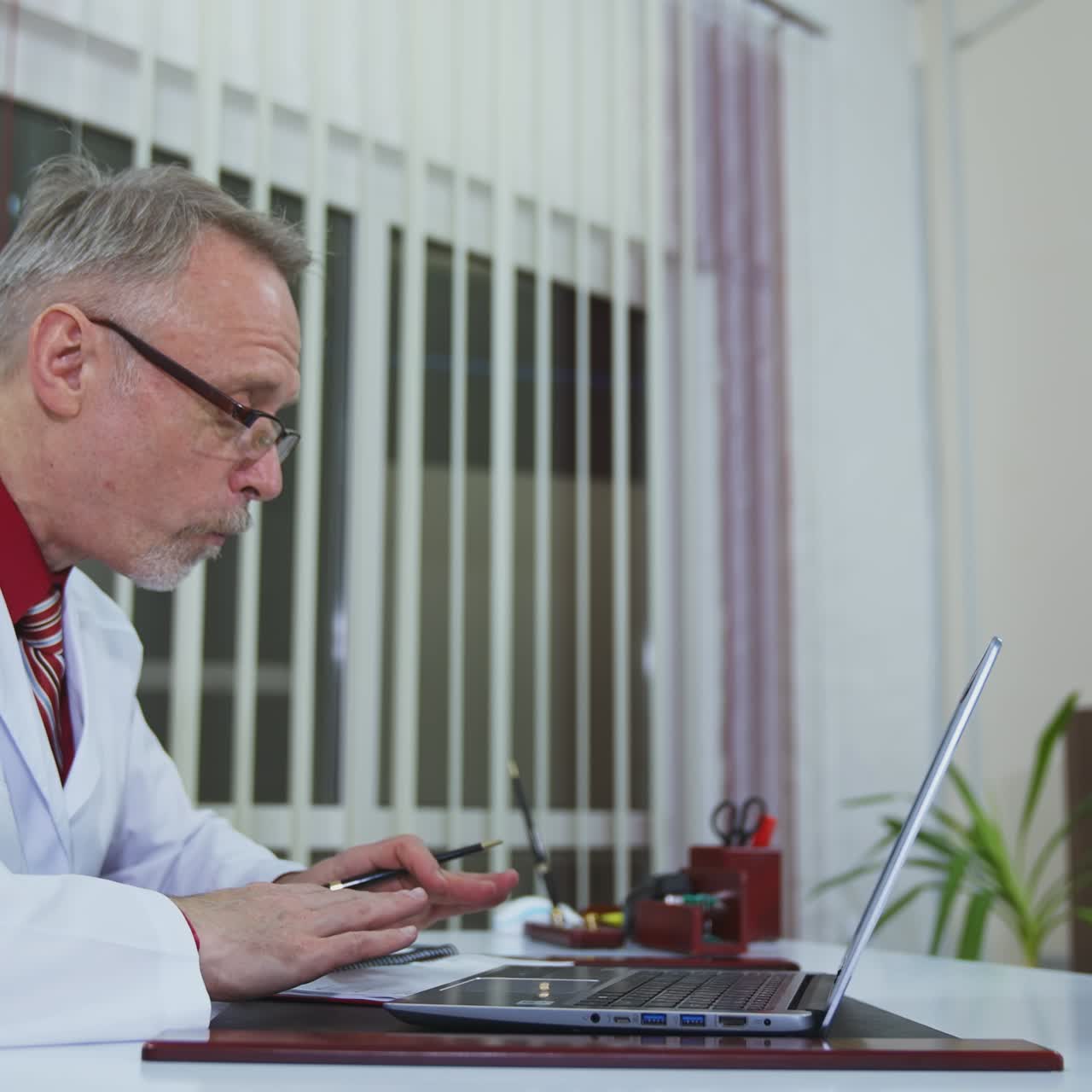Happy doctor working on a laptop in modern clinic. Online consultation. Experienced mature medic in scrubs in clinic speaking with patient online