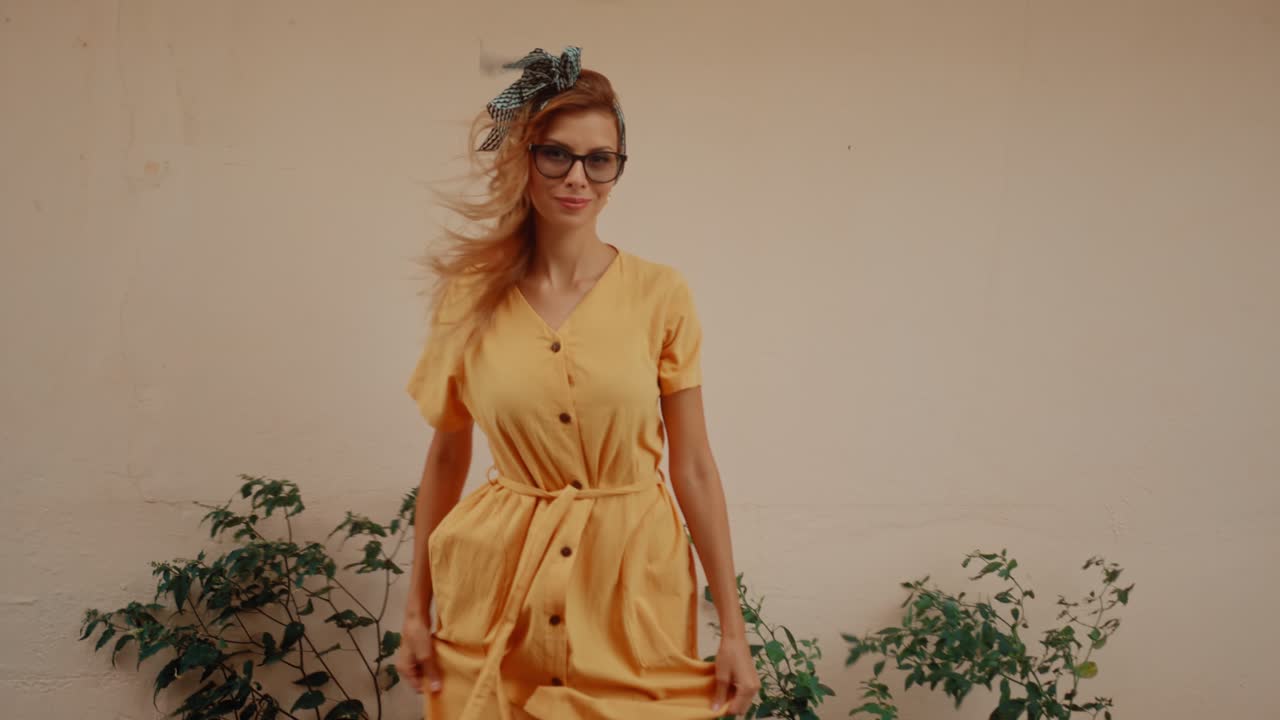 A stylish woman in a yellow dress, glasses, and a headband smiles outdoors
