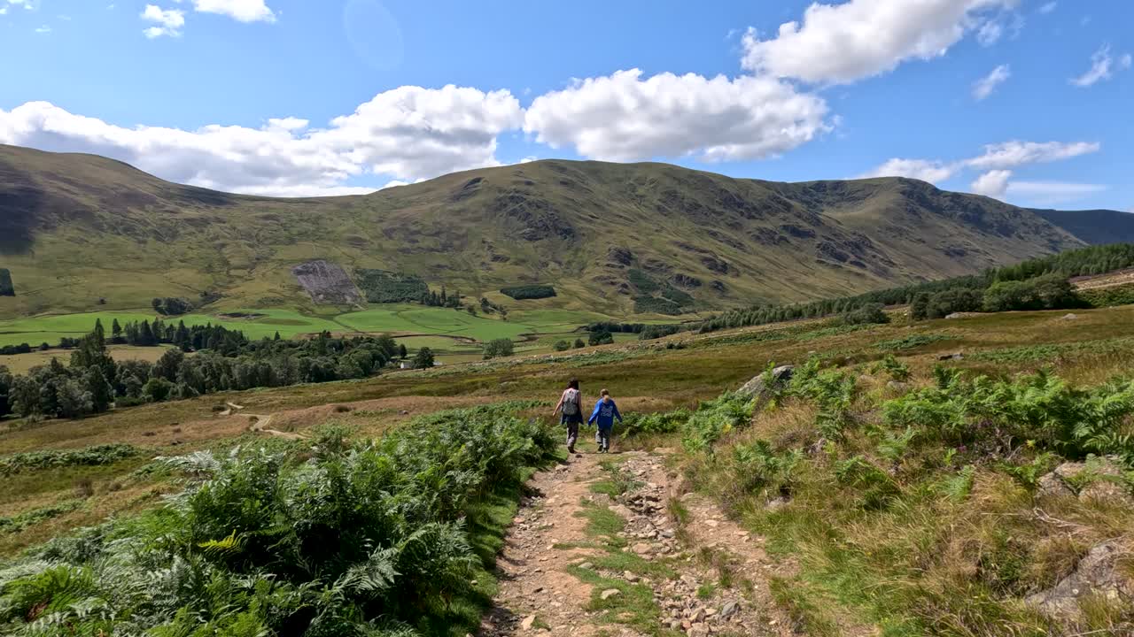 Two hikers walk along a rugged dirt trail through green hills under bright daylight, with wide camera movement capturing the scenic Scottish landscape