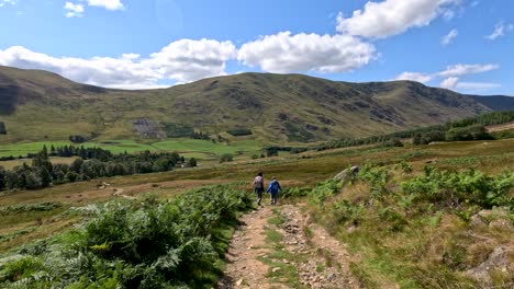 two hikers walk along a rugged dirt trail through green hills under bright daylight, with wide camera movement capturing the scenic scottish landscape