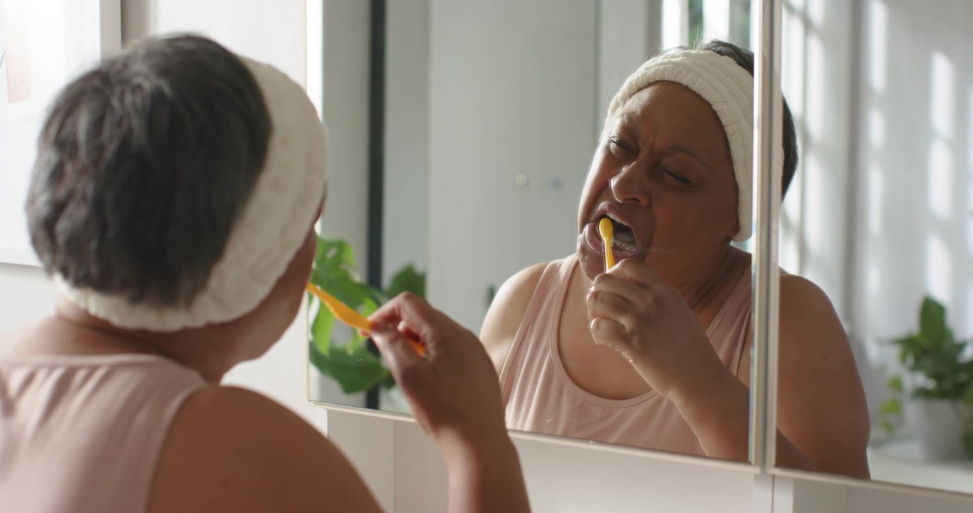 Mature woman bringing yellow toothbrush brushing teeth checking mirror in bathroom for hygiene