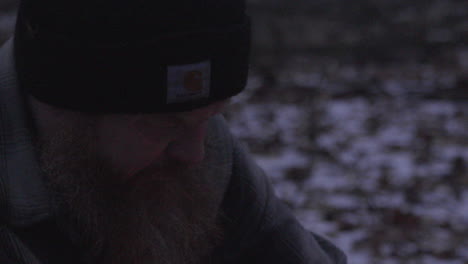 close up of the face of a bearded man in a winter hat is starting a campfire in a wooded area with snow covered fallen leaves in the background and fire lights the mans face