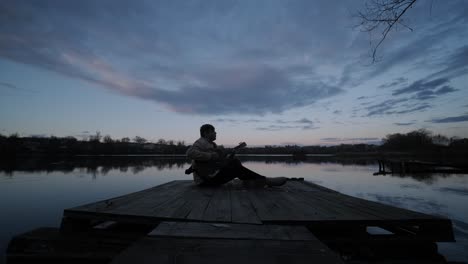 silhouette of musician in black playing guitar sitting on pier embankment on sunset