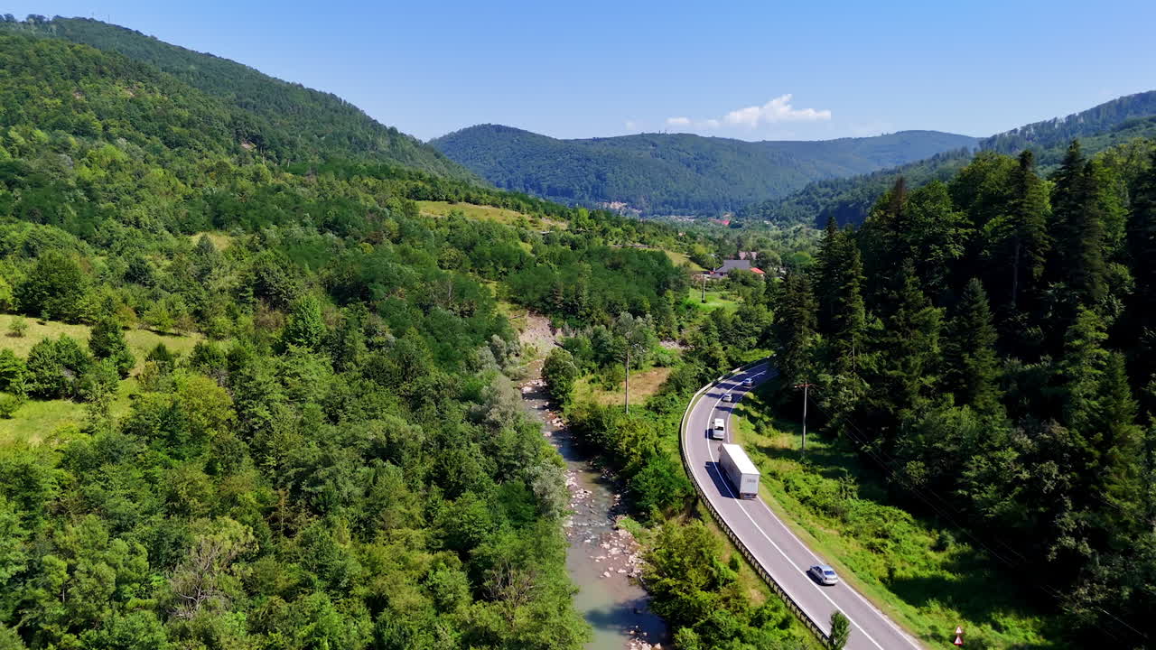 Mountain river and road in a lush Romanian valley. Aerial drone view of a river flowing next to a curving mountain road surrounded by green trees and hills in Romania