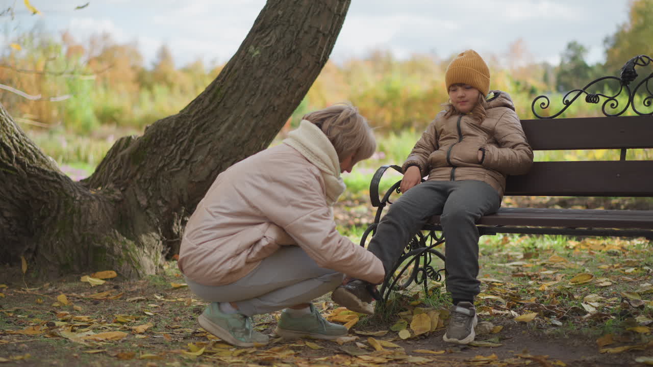 tender moment as mother crouches down to unstrap kid shoe beside wooden bench in serene autumn park setting, fallen leaves scattered across ground beneath leaning tree in peaceful outdoor scene