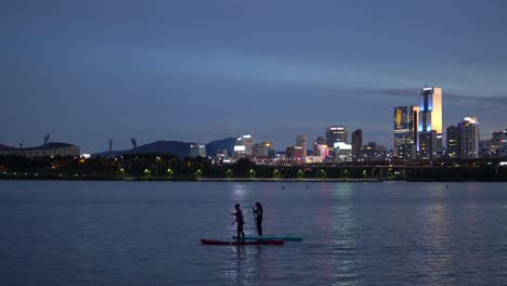 silhouette of two people stand up paddleboarding on han river at twilight, cheongdam bridge with night cars traffic and jamshil district of seoul on colorful purple sky background, static 4k