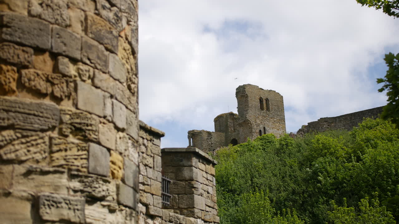 A castle ruin overlooks dense greenery in Scarborough, North Yorkshire, England, with tall stone walls and tower remains under a partly cloudy sky