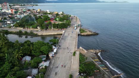 picturesque aerial orbit of a quaint, coastal philippine town, boulevard and bridge with motorcycles driving during daytime