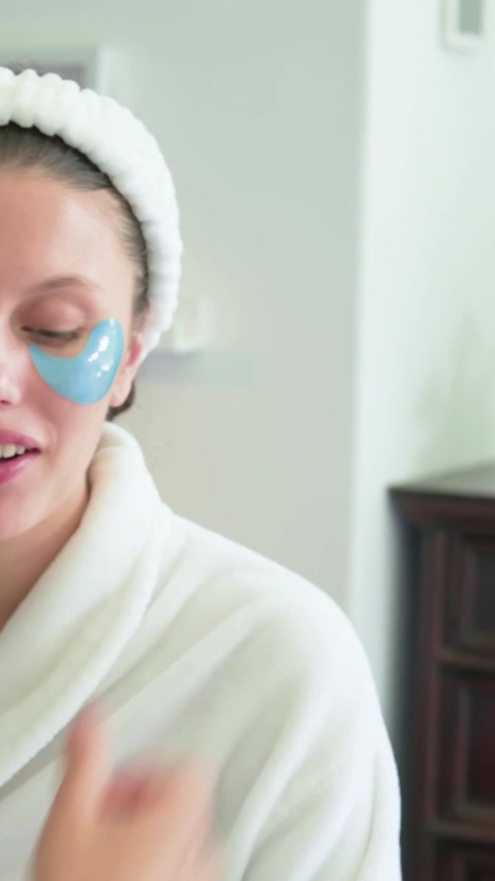 Vertical video: Pressing hand placing blue eye pads onto woman in robe, headband at home for care