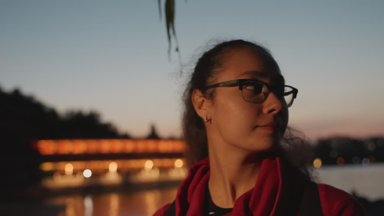 Twilight waterfront portrait of woman gazing, student with backpack, traveler pausing, artist reflecting against softly lit bridge bokeh glasses and red scarf frame profile, long hair catching breeze