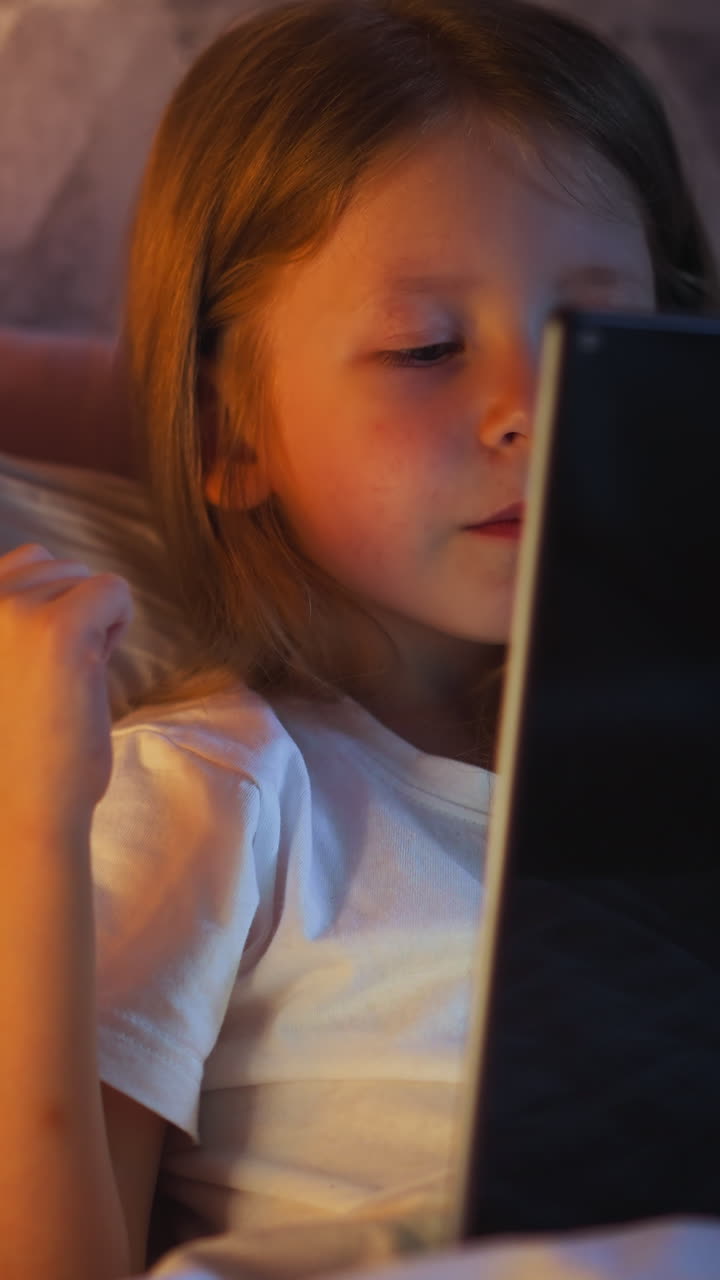 Joyful little girl preschooler happy of winning in videogame expresses joy with raising fist holding modern tablet on bed illuminated by night lamp light in evening slow motion