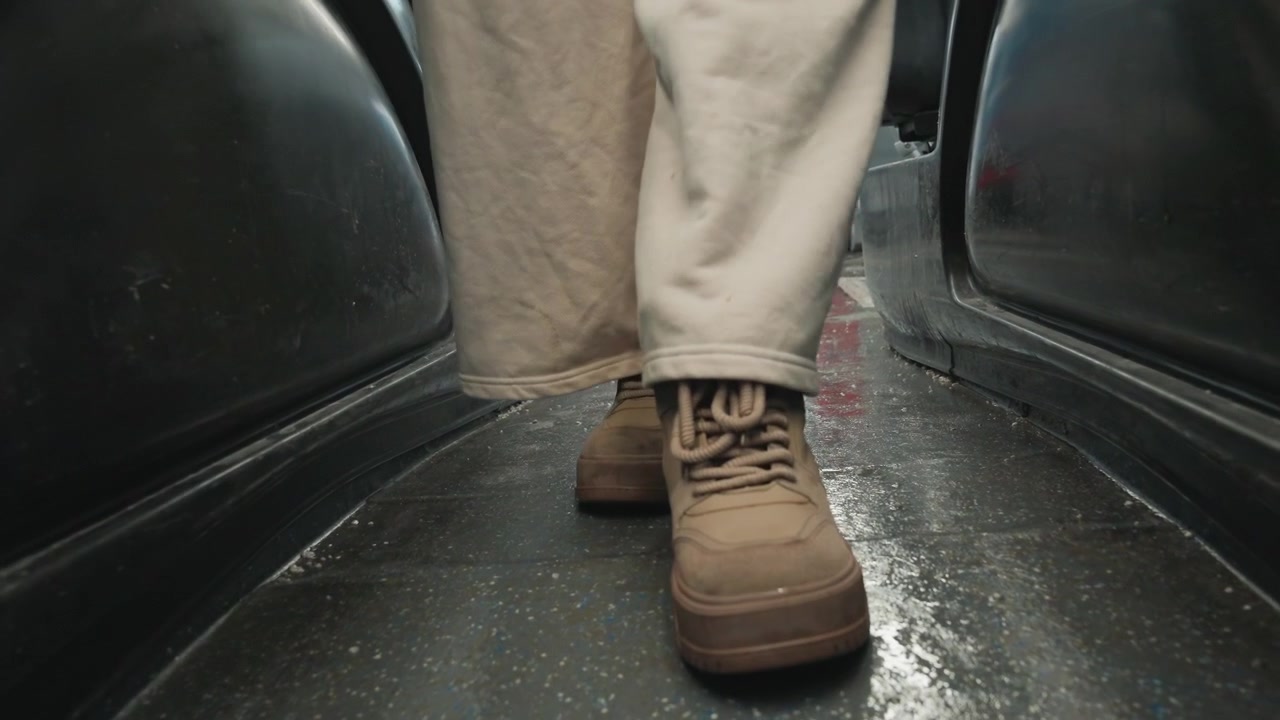 Floor level view of brown lace-up boots and light trousers walking through narrow public bus aisle; visible motion shows planted step, lifted foot, worn soles, scuffed texture, and steady travel