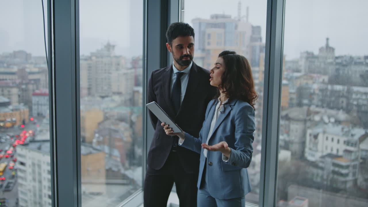 Business partners speaking standing at office window looking on city view.