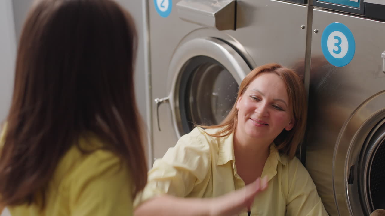 Housewife seated by washer loads fabrics while school girl helper swings towel, shares high five, teamwork in modern laundromat, stainless machines, joyful routine, close view shows cooperation