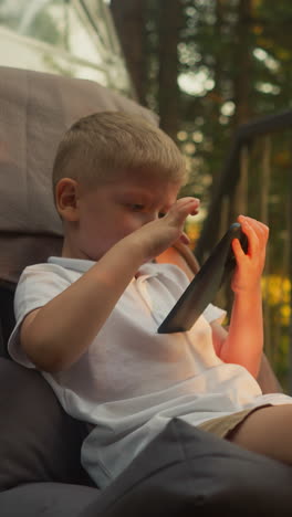 little boy plays with smartphone sitting on balcony in glamping house surrounded by dense forest. sun rays fall on playful child having rest slow motion