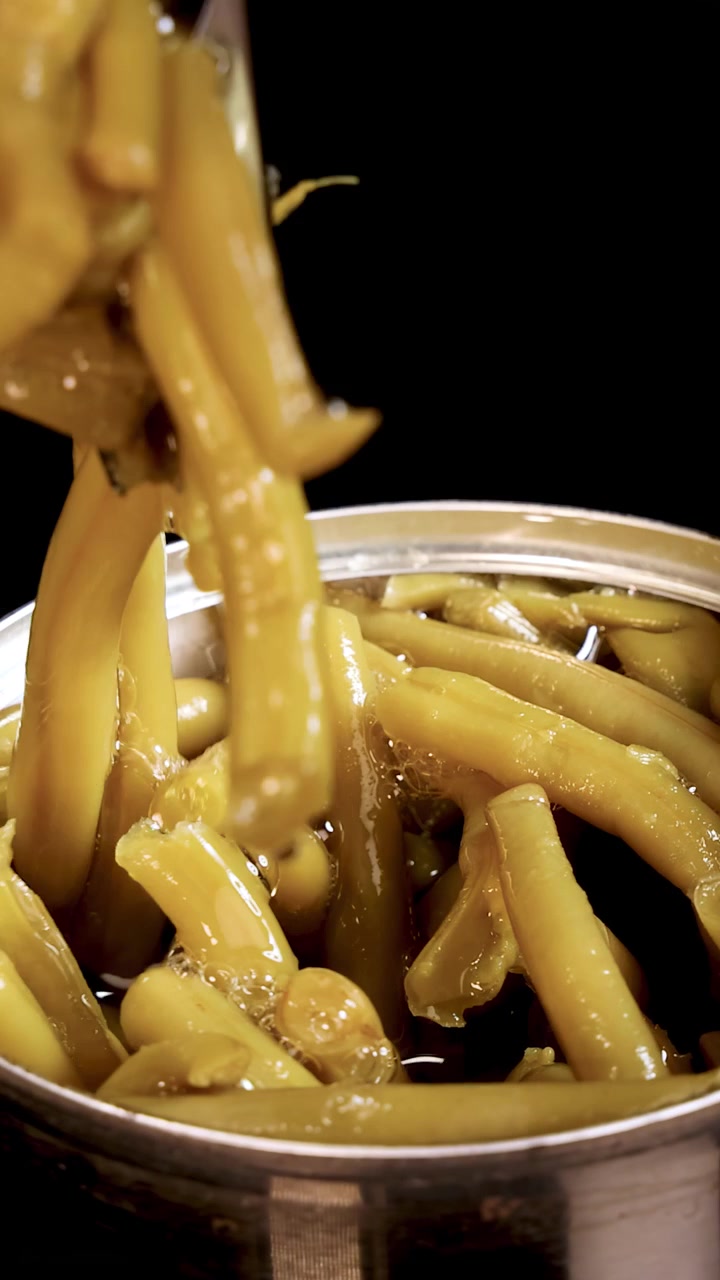 A hand uses metal tongs to pick up cooked haricot beans from an opened canned food container under bright, direct lighting with a black background