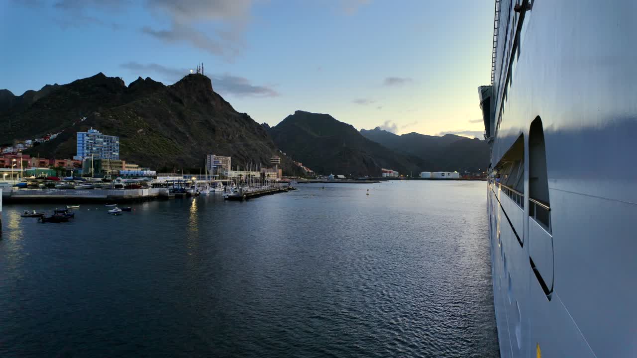 View from a cruise ship docked at Santa Cruz de Tenerife. Marina, city buildings, and Anaga Mountains are visible under warm evening light with calm sea reflections.