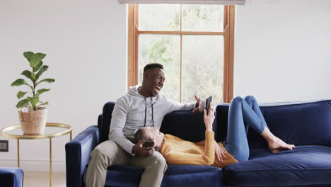 Happy-diverse-couple-sitting-and-lying-on-sofa-and-using-smartphones-in-home,copy-space