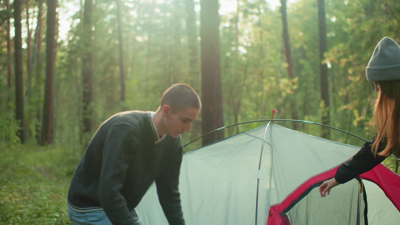 young tourists adjust camping tent in forest as setup nears completion surrounded by tall trees and nature during peaceful outdoor adventure with hints of teamwork and early morning light