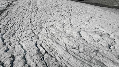 Aerial-flyover-around-a-hiker-walking-across-the-ice-of-the-Allalin-glacier-near-Saas-Fee-in-Valais,-Switzerland-on-a-sunny-day-in-the-Swiss-Alps