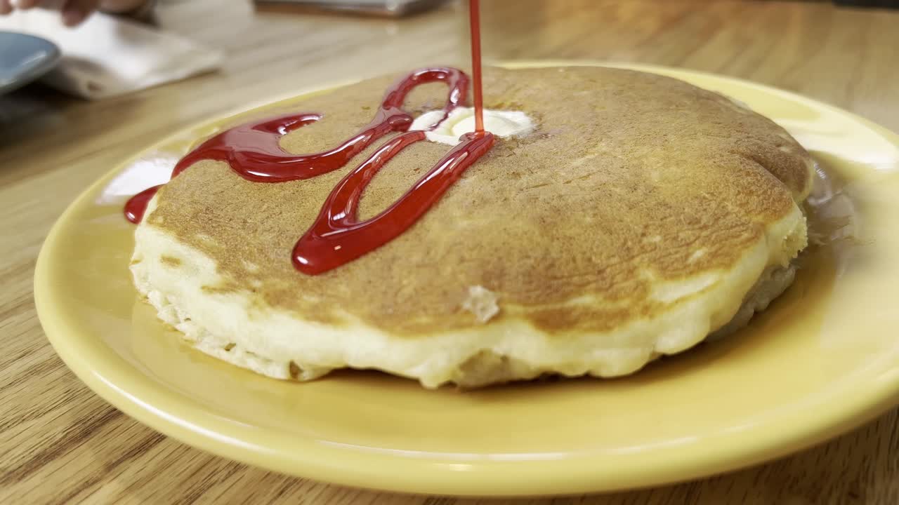 Cook pouring rich strawberry syrup over a stack of fluffy pancakes topped with creamy butter, served on a vibrant yellow plate, creating a delightful sweet breakfast experience