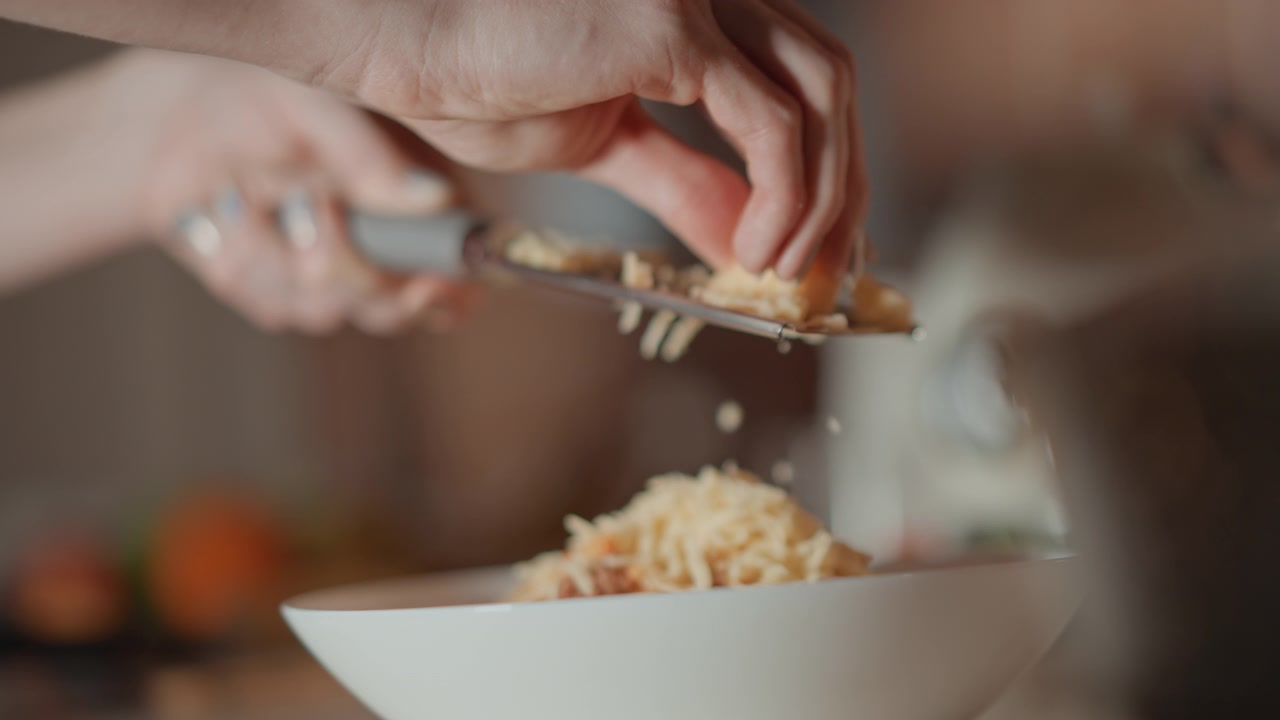 Mani che premono un blocco di formaggio su una grattugia di metallo mentre stabilizzano la grattugia sopra una ciotola bianca, movimento verso il basso visibile che produce scaglie sottili che cadono nella ciotola.