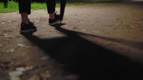 close up of lady legs walking on paved path holding umbrella, scattered dry foliage on ground, dim lighting creating long shadows, greenery visible in background