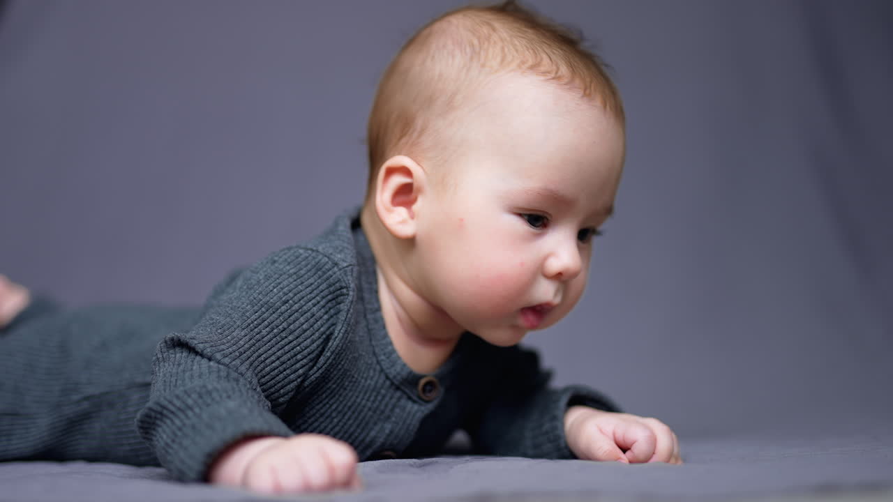 Few months old toddler learned how to keep head steady. Curious kid looks around lying on bed. Grey blurred backdrop.