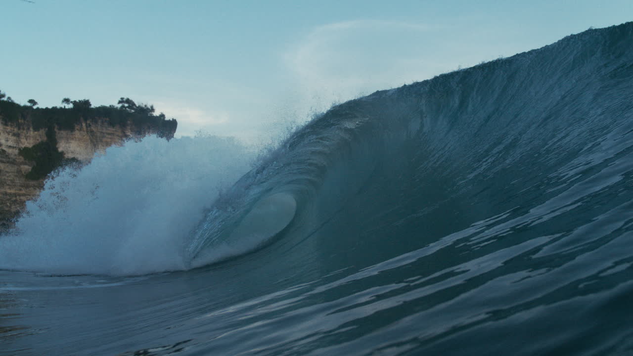 Clean wave curls toward rocky shore under soft afternoon light in tropics, barreling with eruption of foam