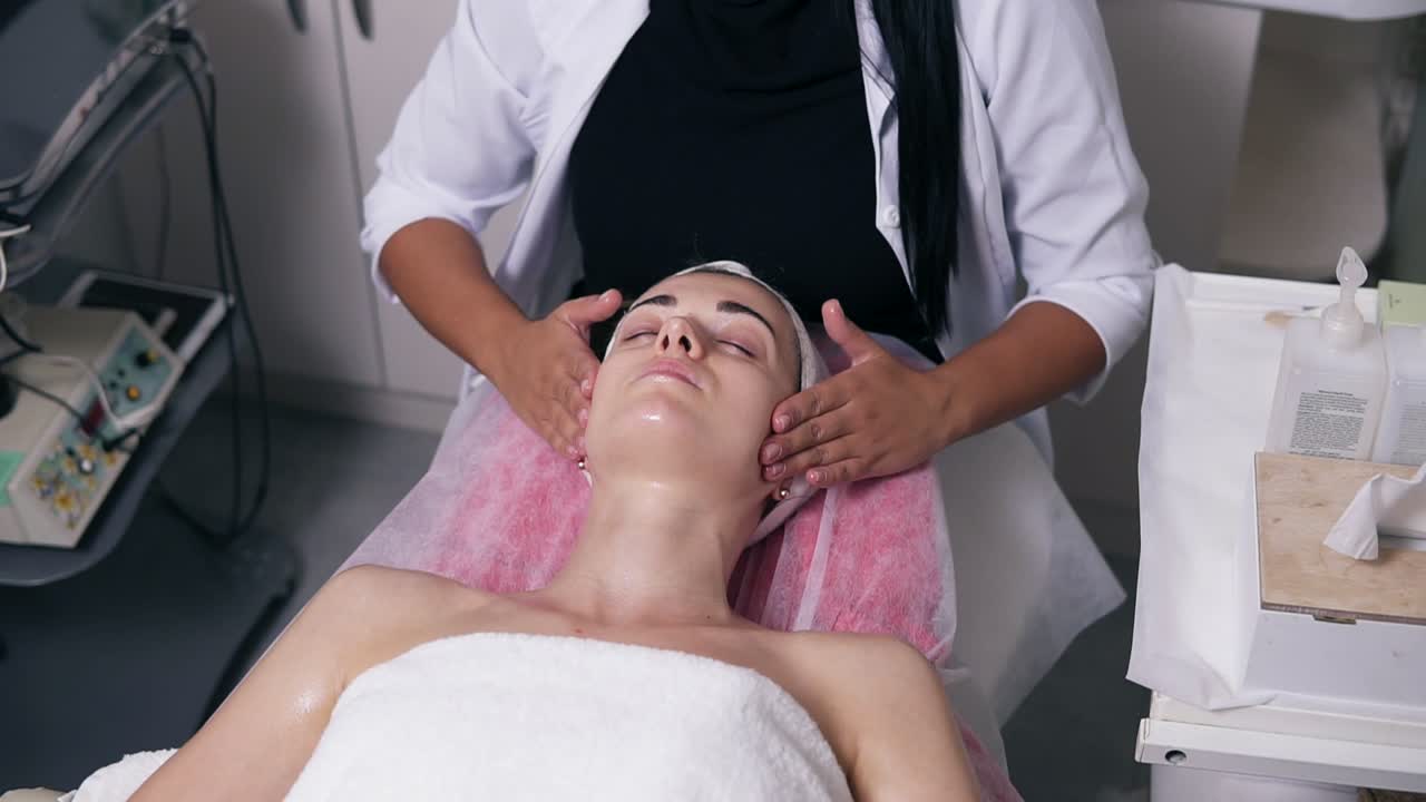 la cosmetóloga está haciendo un masaje facial en el salón de spa. una mujer joven con los ojos cerrados está acostada en el sofá.