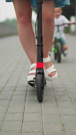 una mujer monta un scooter negro en una pasarela pavimentada, sus piernas visibles mientras se mueve hacia adelante, un niño la sigue de cerca en una pequeña bicicleta