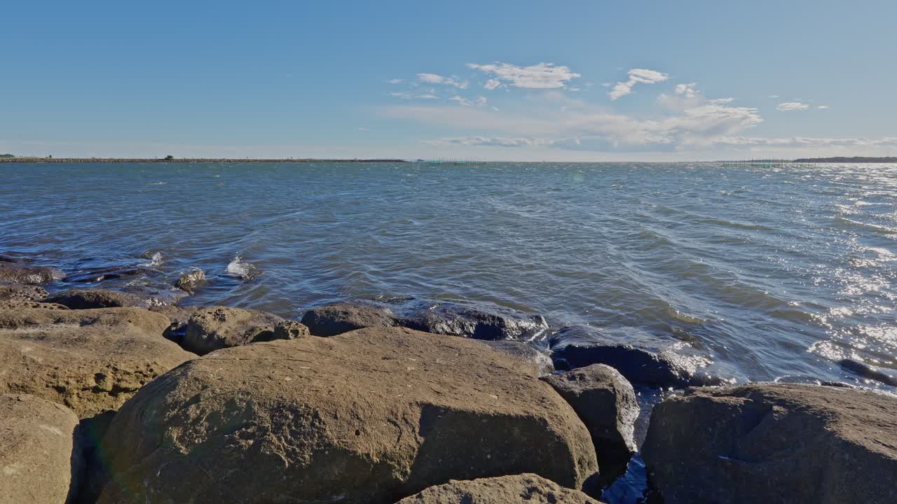 A wide shot of the vast, choppy water of Tokyo Bay, framed by large, dark rocks in the foreground under a bright sky