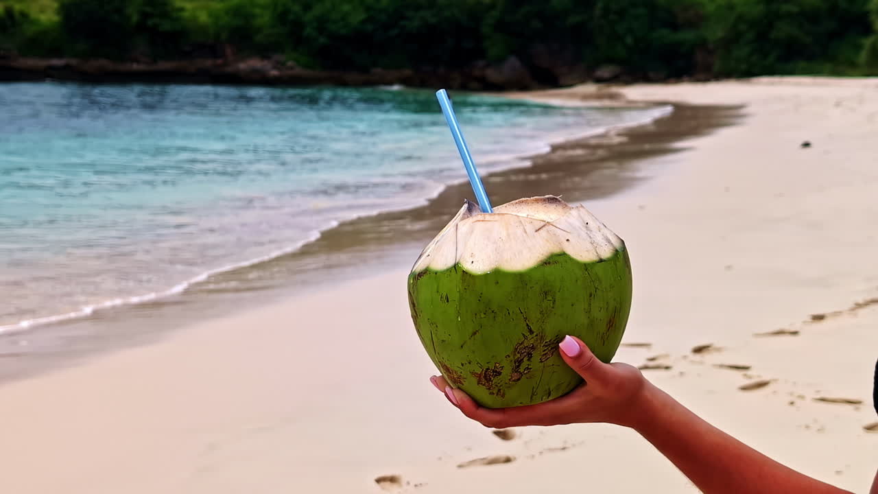 A woman's hand with pink nails holds a fresh green coconut drink with a straw, relaxing on the famous Pink Beach with turquoise water in Lombok, Indonesia, on a summer day