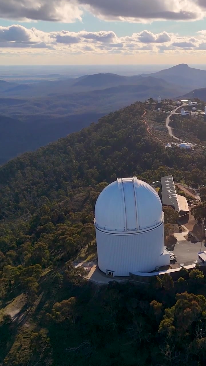 Um drone desliza sobre uma enorme cúpula de observatório astronômico situada em um cume de montanha arborizado.