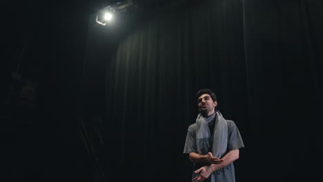 Un-Hombre-Moreno-Seguro-De-Sí-Mismo-Con-Una-Camiseta-Gris-Y-Una-Bufanda,-El-Director-De-Escena,-Sube-Al-Escenario-Y-Saluda-A-Todos-En-El-Teatro-Con-Cortinas-Negras.