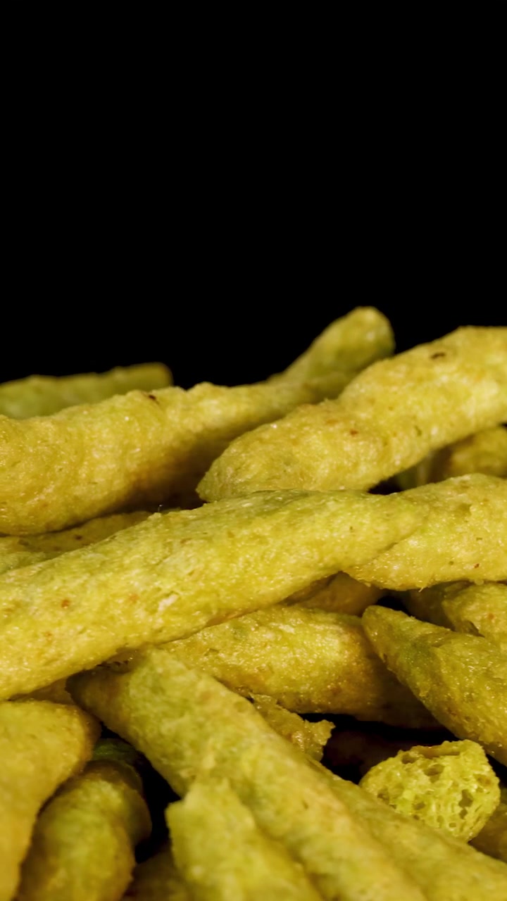 Macro rotating view of green pea puffed snack sticks against black background, with dramatic lighting