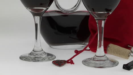 glasses filled with red wine and surrounded by a halffull carafe and red velvet bag are displayed on a white surface