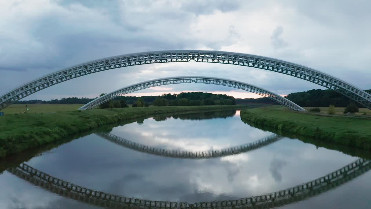 Flying under two bridges for gas pipes running across the calm river Moravia on the border of Slovakia and Czechia. Aerial sunset footage of two symmetrical gas pipe bridges reflecting in water