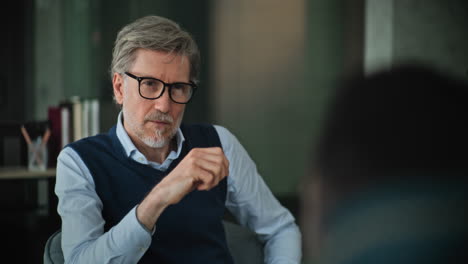 a man in a blue shirt and glasses is sitting in a chair and listening to someone. he is serious and thoughtful.