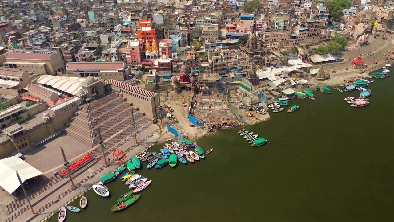 Una ripresa aerea ampia e statica che mostra lo storico Manikarnika Ghat, templi indù tradizionali e barche di legno colorate ormeggiate lungo il sacro fiume Gange a Varanasi, India.
