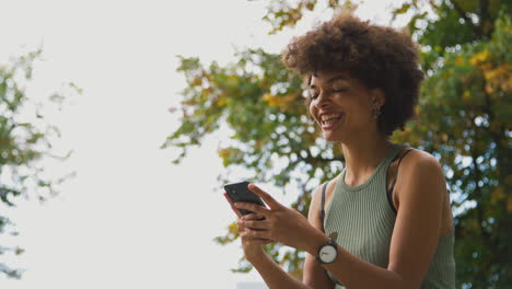 Lächelnde-Junge-Frau-Im-Freien,-Die-Lacht,-Während-Sie-Nachrichten-Auf-Dem-Mobiltelefon-Sendet