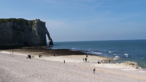 tomada panorámica de personas caminando en la playa de guijarros de etretat con la famosa formación de acantilados marinos en el fondo - hermoso día soleado con un cielo azul claro en francia
