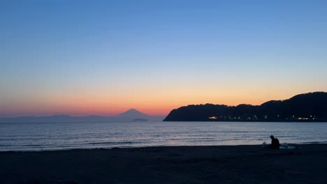 Couple-sitting-on-a-beach-at-dusk-with-a-view-of-distant-mountains-and-vibrant-sunset-sky