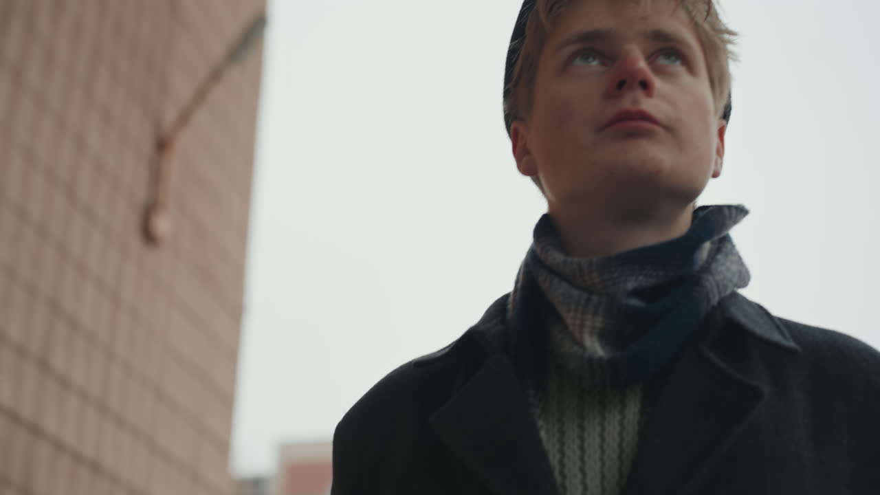 Young caucasian creator looking up sky, lowangle rainy portrait, resolve in expression, droplets on beret, scarf around neck, city facade background, cinematic hope, directorial focus on future scene