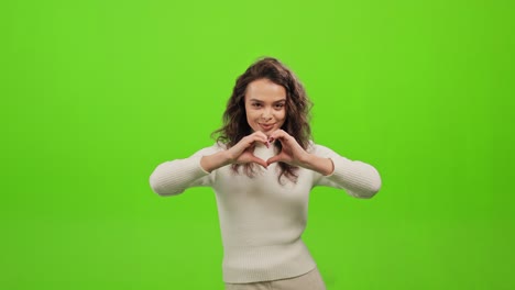 la mujer está de pie y mirando a la cámara. está poniendo sus manos juntas en forma de corazón. está de pie sobre un fondo verde. pantalla verde. 4k.
